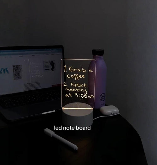 LED note board on a desk with a laptop and water bottle in a dimly lit room.